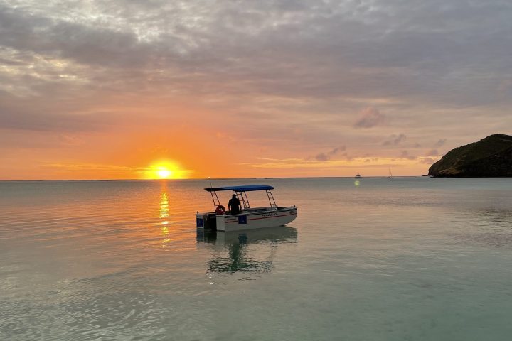 Small boat on calm sea at sunset with cloudy sky and hill in background.