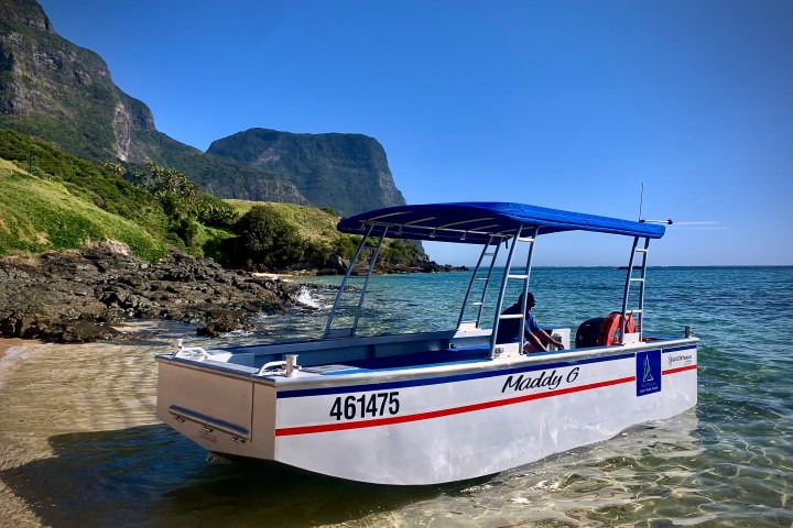 Boat named 'Maddy G' on clear water near rocky shore and green hills under blue sky.