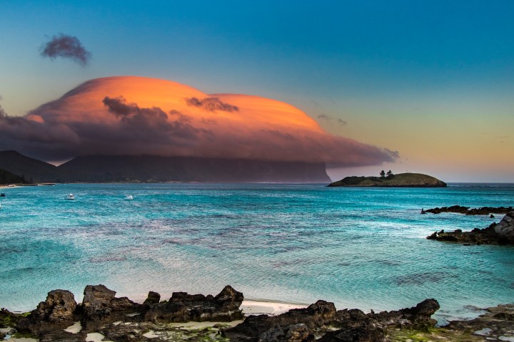 Dramatic orange cloud over island at sunset with boats on turquoise sea.